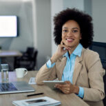 Happy African American businesswoman working on touchpad in the Happy black entrepreneur working on wireless technology at her office desk and looking at camera.